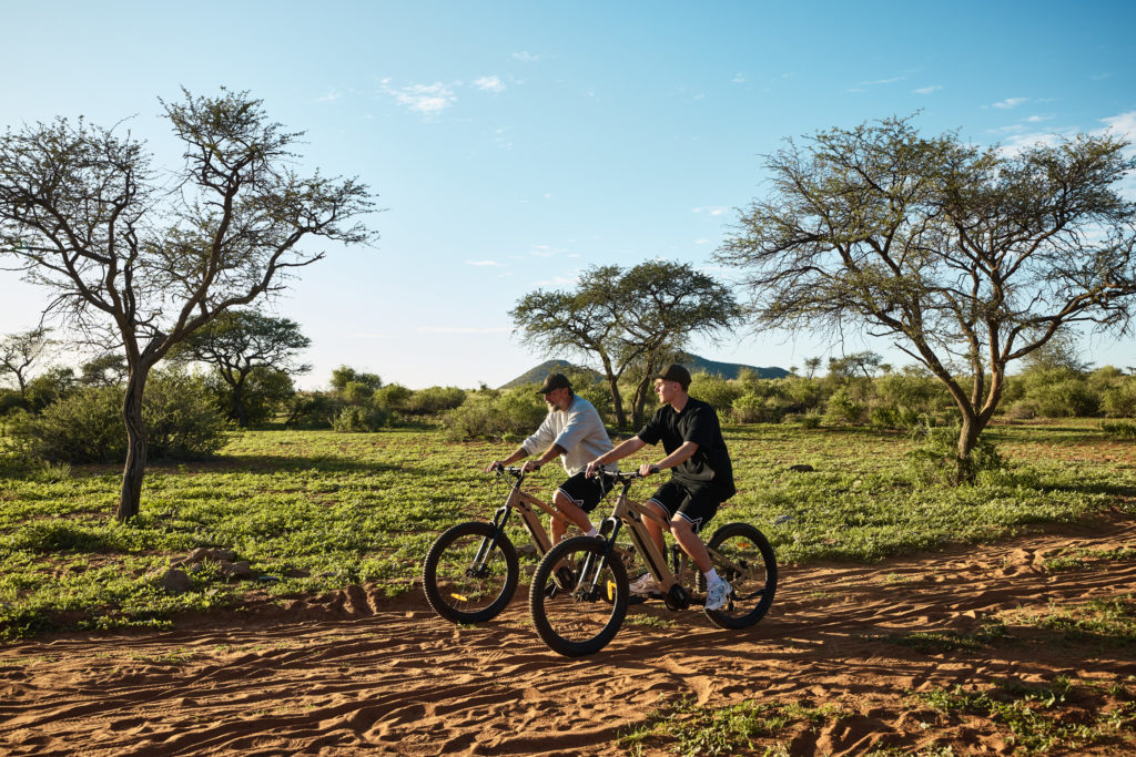 Two guests ride e-bikes along a sandy trail beneath acacia trees