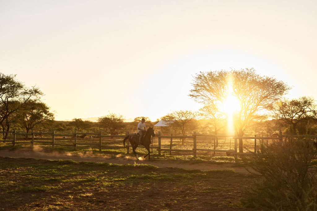 A guest rides on horseback through the paddocks as the sun sets over the Kalahari