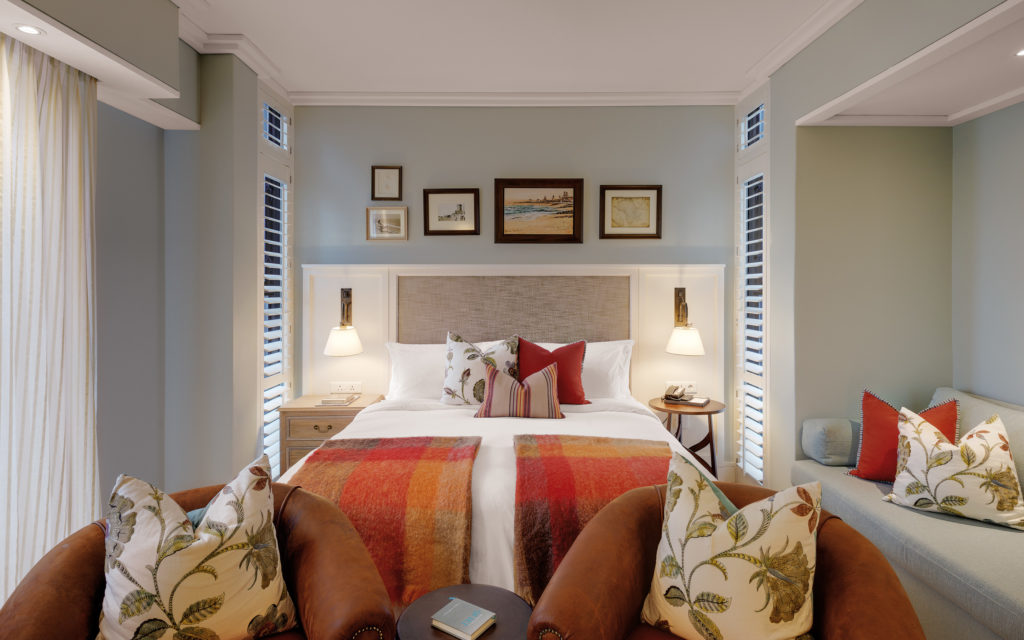 Luxury Room at Strand Hotel Swakopmund with tan leather armchairs, a red and orange plaid throw and a gallery wall of framed coastal artwork above the bed