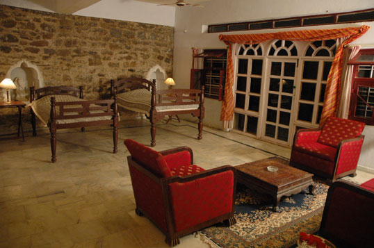 A stone-walled sitting room at Darbargadh Poshina with carved wooden day beds, red upholstered armchairs and arched shuttered windows