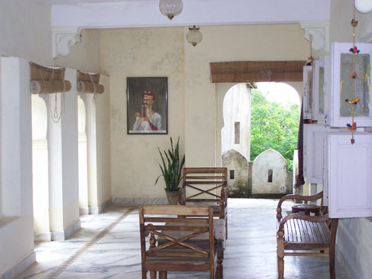 A shaded verandah at Darbargadh Poshina with teak furniture, a framed royal portrait and views through arched doorways to the garden