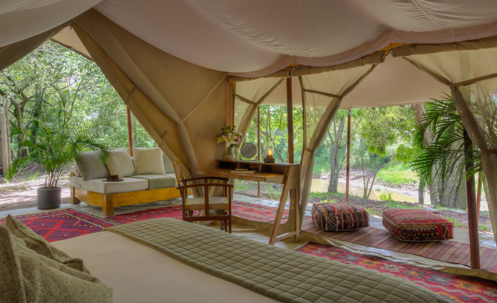 Inside a Naibor double tent, a daybed and writing desk sit beside the open canvas doorway, with kilim rugs underfoot and a view through to the veranda and river beyond.
