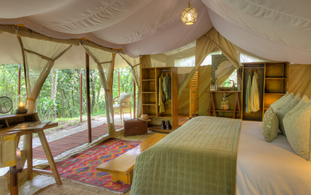 A Naibor tent interior shows an open wardrobe, wash basin and hanging lantern beside the bed, with a suitcase and boots set out near the entrance.