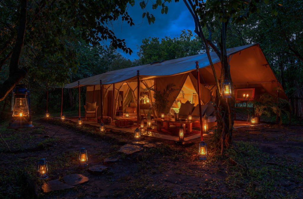 A Naibor guest tent glows warmly at dusk, its veranda lined with lit lanterns leading along a wooden walkway through the trees.