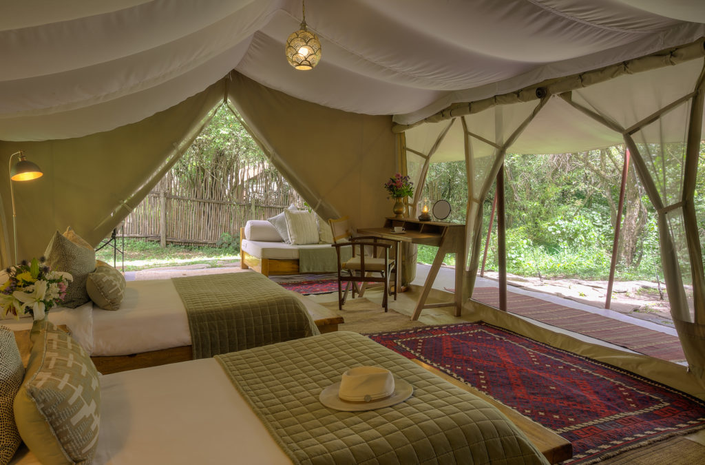 A twin-bedded Naibor tent is furnished with green quilted bedspreads, a writing desk and floor lamp, opening onto a veranda framed by riverine forest.