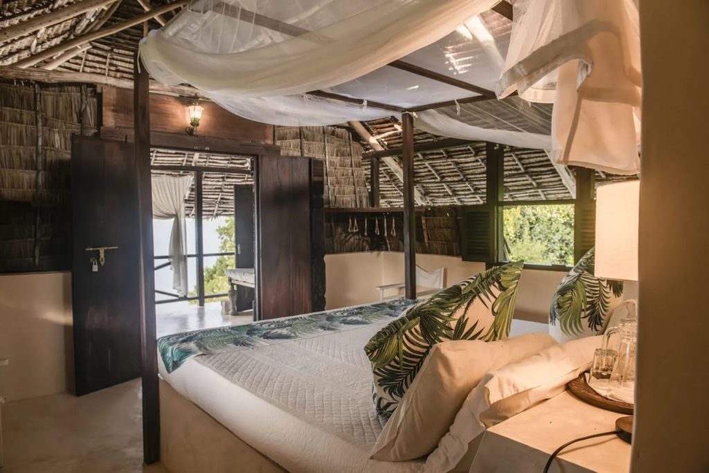 Four-poster bed with draped mosquito net and palm-print cushions under a thatched roof, doors opening to a shaded veranda.