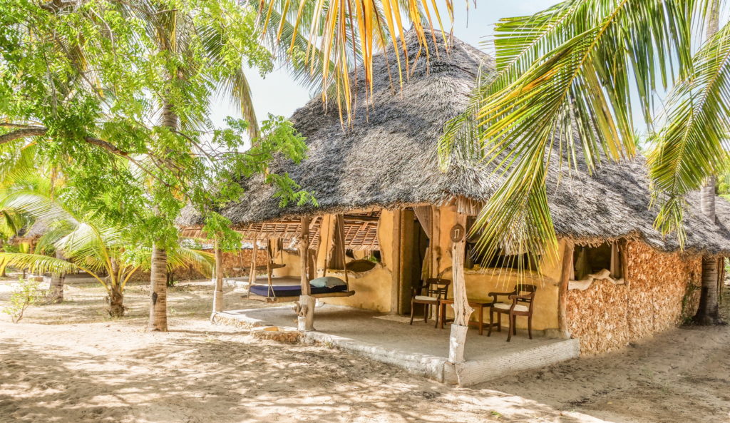 Thatched cottage 17 with a coral stone wall, rope swing daybed and wooden chairs on its veranda, shaded by palms at Manda Bay Lodge.
