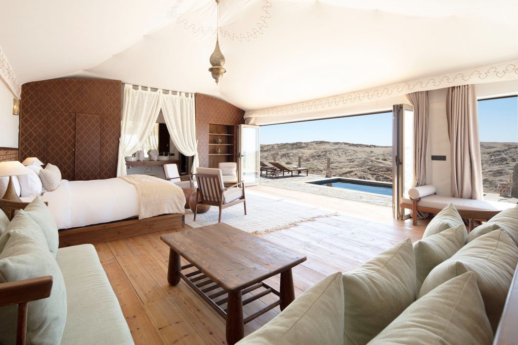 A suite bedroom with sage-green sofas and a wall of glass opening onto a plunge pool and desert horizon.
