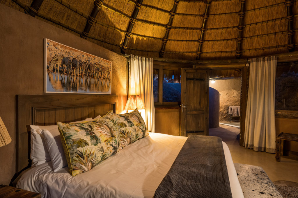 Firelight and lamplight warm a thatched Mowani suite by night, a zebra print above the bed and the open-air bathroom glimpsed beyond the door.