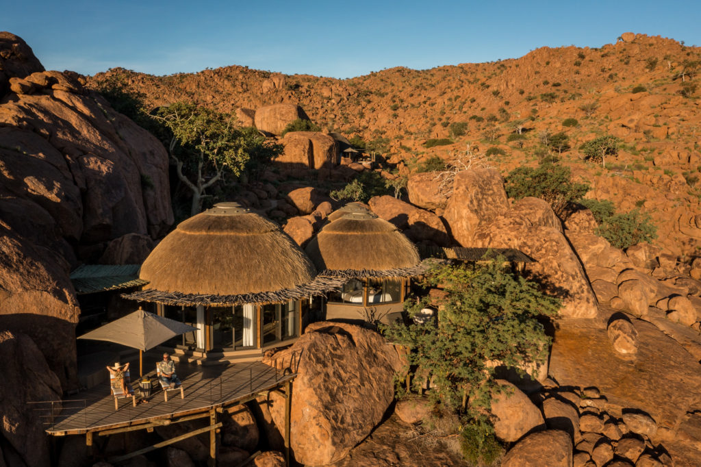 Two guests relax on a boulder-framed deck outside Mowani's glass-walled Mountain Suite as the last sun catches the surrounding rock.