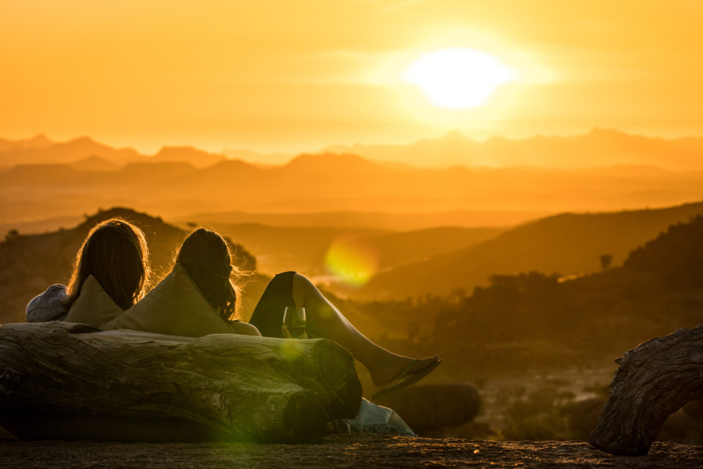 Two guests lie back with wine in hand at sundowner hour, the Damaraland hills fading into layers of gold and amber light.