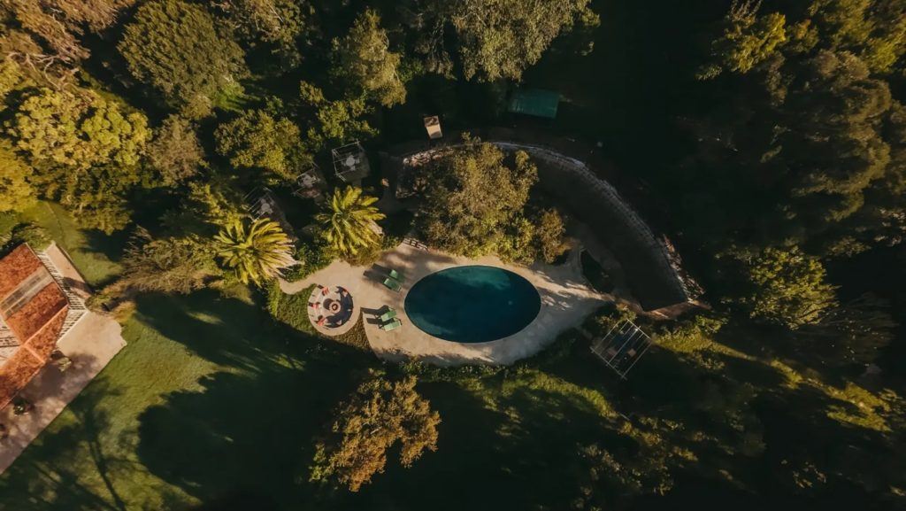 Aerial view of a round swimming pool, fire pit and loungers surrounded by dense forest and lawn in late afternoon light.