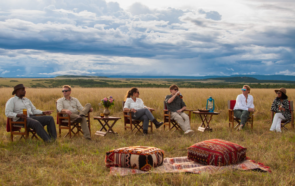 Six guests and their guide relax with drinks in canvas chairs on the open plains as a storm gathers on the horizon during a bush sundowner.