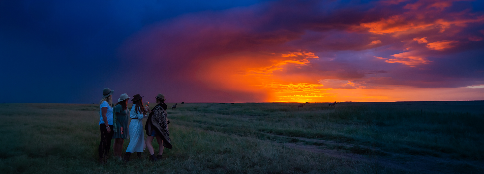 Four guests stand silhouetted against a vivid orange and indigo sunset over the Maasai Mara plains, wildlife visible grazing in the distance.
