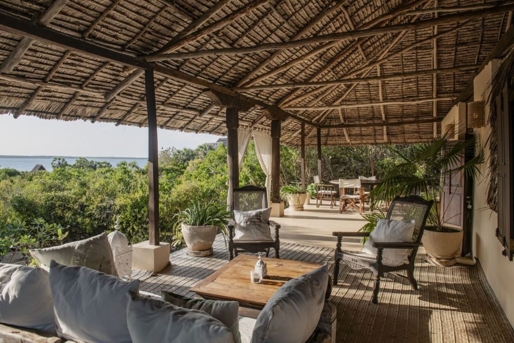 Long thatched veranda with cane armchairs, a cushioned daybed and a dining table beside curtained carved pillars, with sea views.