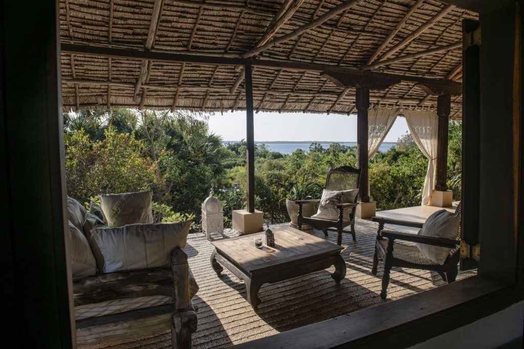 Thatched veranda with cane chairs, a carved coffee table and white lantern looking over coastal bush to the sea at The Cabanas Lamu.