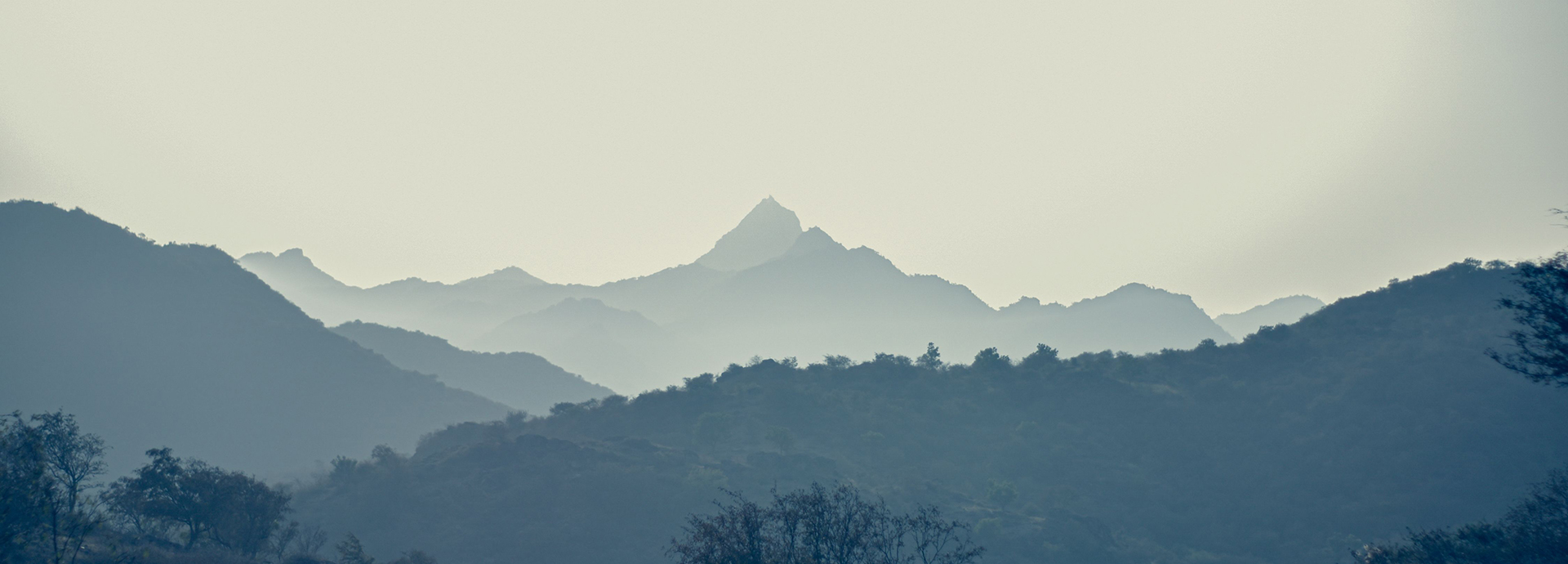 Layered peaks of the Aravalli hills fade into haze beyond Poshina, with a distinctive pointed summit rising above the ridgeline