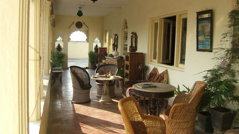 A sunlit verandah at Darbargadh Poshina lined with wicker chairs, antique wooden furniture and views through an arched doorway