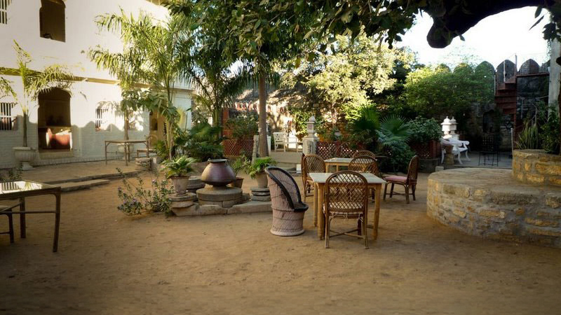 A shaded courtyard garden at Darbargadh Poshina with rattan seating, potted palms and an old stone well beneath spreading trees