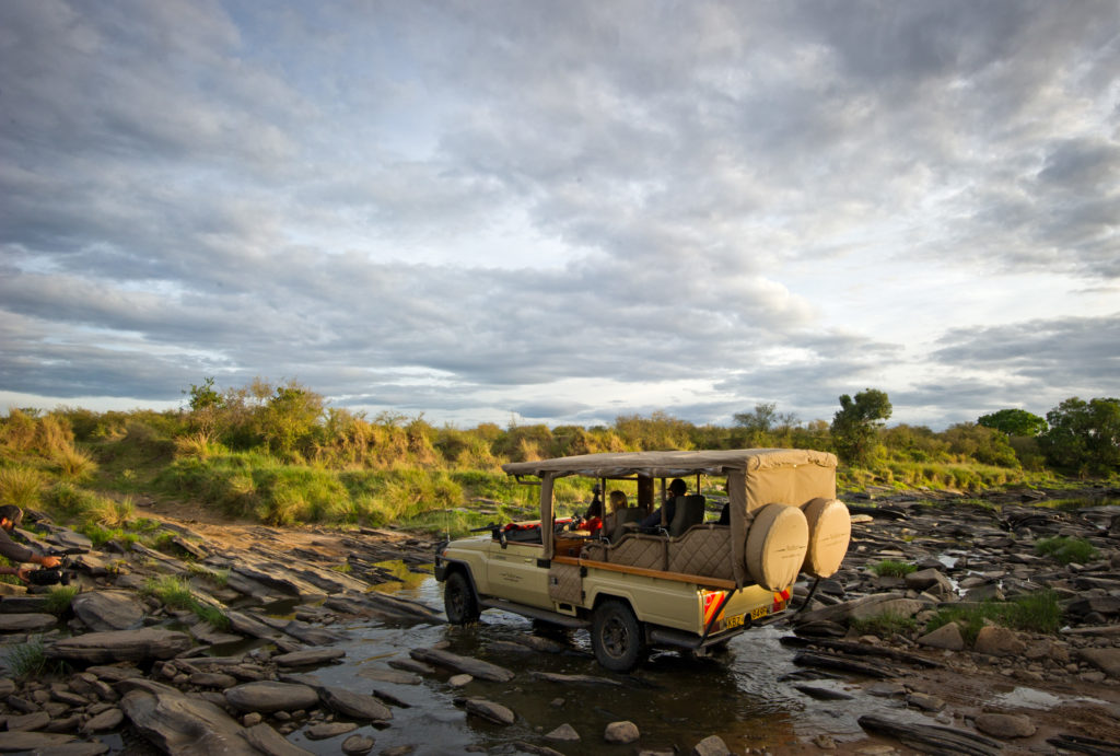 A Naibor safari vehicle fords a shallow, rock-strewn stretch of river during a game drive, with a guest filming the crossing from the bank.
