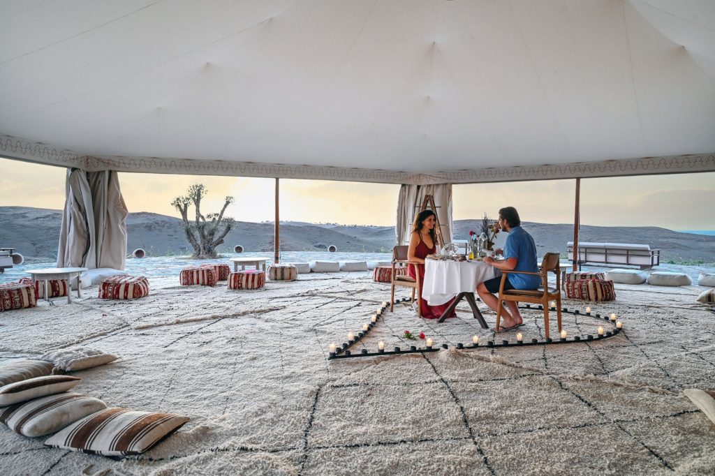 A couple share a candlelit dinner beneath an open-sided tent, the desert stretching out behind them.