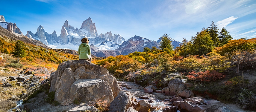 A hiker seated on a boulder gazing towards Mount Fitz Roy, surrounded by autumn colours in Argentina’s Los Glaciares National Park.