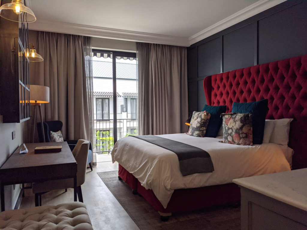 Double room at The Weinberg Windhoek with deep red tufted headboard, patterned cushions and balcony view over the estate