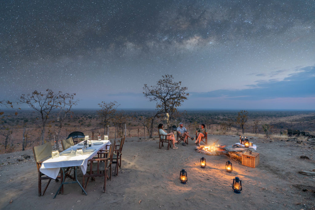 Guests dine and gather around a fire beneath a starlit sky on a bush sleepout in the Lower Zambezi.