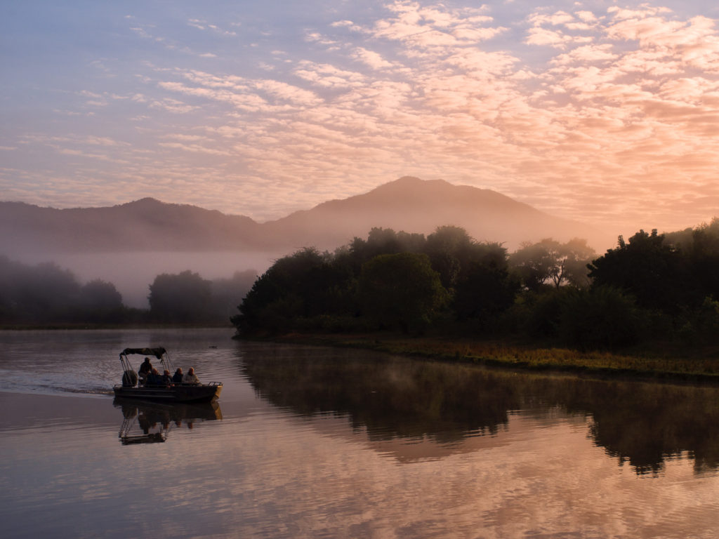 A boat carries guests along the misty Zambezi at dawn, mountains rising beyond the shoreline.