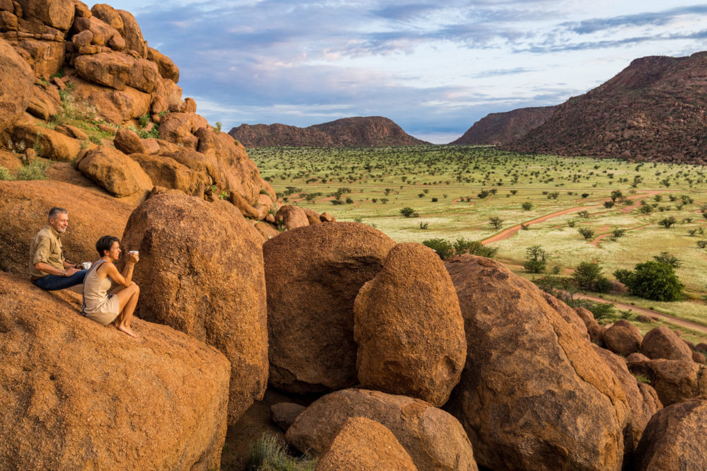 A couple shares coffee atop weathered boulders overlooking a green Damaraland valley, tracing the walking trail's viewpoint.